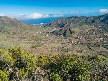 Lush green valley with terraced fields and village Las Portelas. Landscape with rocks and hills seen from hiking trail at Park rural de Teno, Tenerife, Canary Islands, Spain. sunny day, blue sky.