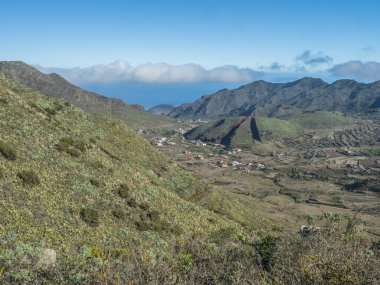 Lush green valley with terraced fields and village Las Portelas. Landscape with rocks and hills seen from hiking trail at Park rural de Teno, Tenerife, Canary Islands, Spain. sunny day, blue sky.