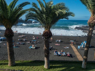 Santiago del Teide, Tenerife, Canary Islands, Spain, december 24, 2021: View of black sand beach Playa de la Arena with sunbathing and swimming tourist people and palm trees at sunny christmas day.