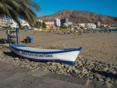 Los Cristianos, Tenerife, Kanarya Adaları, İspanya, 2 Ocak 2022: Playa de Los Cristianos 'un kumsalında mavi ahşap teknesi, güneşlenen turistler, verimsiz tepeler ve apartmanlar.