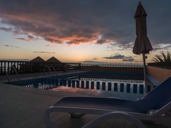 stock image View of a sun bed and umbrella at pool area on rooftop of apartment in Valle Gran Rey during red orange sunset reflecting on the water. La Gomera, Canary Islands, Spain. Luxury holiday concept-