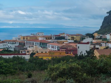 Kuzey kıyısındaki yeşil vadide renkli geleneksel evleri, tarlaları ve okyanusu olan eski Agulo köyü manzarası. Bulutlu bir kış günü. La Gomera, Kanarya Adaları, İspanya.