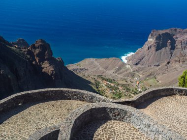 Palmiye ağaçları, kaktüsleri ve La Merica dağının keskin uçurumları olan yeşil vadi manzarası. Camino La Merica yürüyüş parkurunda görülmüş. Arure, Valle Gran Rey, La Gomera, Kanarya Adaları, İspanya