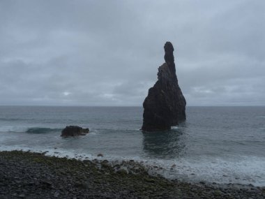 Porto Moniz yakınlarındaki Madeira Adası 'nın kuzey kıyısındaki kayalık plajda Ilheus da Ribeira da Janela kayalık oluşumu görülüyor. Denizde üç yüksek kaya. Tek renkli, yağmurlu bulutlu bir gün.