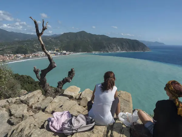 Arkadan iki kadın turist Punta Manara 'daki Torre Saracena' dan Riva Trigoso 'nun hava manzarasını izliyorlar. Ön planda kayalar ve kuru ağaçlar, evler, tersane, Sestri Levante, Liguria, İtalya.