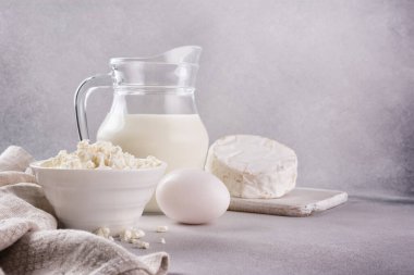 Beyaz kâseli süzme peynir, cam süt sürahisi, tavuk yumurtası ve camembert peyniri. Gri beton arka planda lezzetli bir yemek malzemesi olarak.