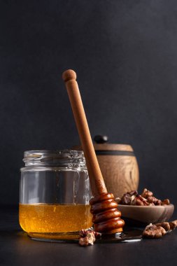 Kitchen composition of wooden barrel and glass jar with healthy organic honey, wooden dipper and bowl of walnuts on dark background