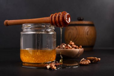Kitchen composition of wooden barrel and glass jar with healthy organic honey, wooden dipper and bowl of walnuts on dark background