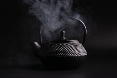 Traditional black asian cast iron hot teapot or kettle with steam on dark background
