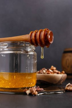 Kitchen composition of glass jar and wooden barrel with healthy organic honey, wooden dipper and bowl of walnuts on dark background