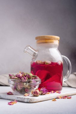 Beautiful flower pink tea in glass teapot with dry rose buds in bowl on grey kitchen background