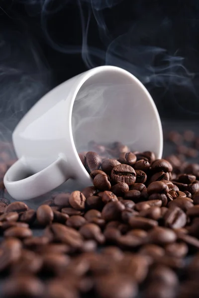 White coffee cup with steam and roasted coffee beans on dark black background