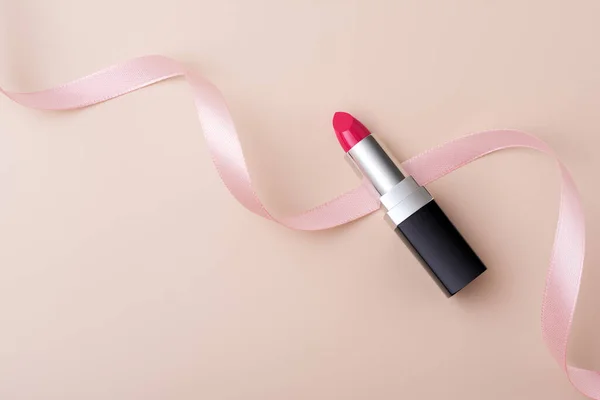 Pink lipstick in black and metal tube packaging with pink decorated ribbon on beige background. Makeup and cosmetics mock-up product concept.