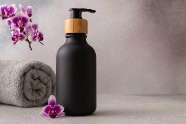 Ahşap bir pompa kapağı olan siyah bir mat şişesi gri bir arka fona doğru merkezden yerleştirilir. Sarılmış gri bir havlu ve canlı mor bir orkide çiçeği görüntünün spa benzeri atmosferini vurgular. Sahne, lüks ve sükunet hissi uyandırıyor.
