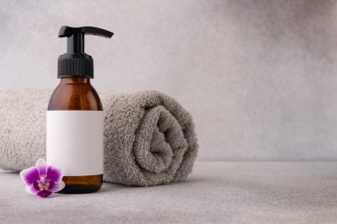 Üzerinde kahverengi bir pompa şişesi ve boş bir etiket bulunan minimalist bir spa düzeneği, yanında arka planda rulo havluyla desenli gri bir yüzeye yerleştirilmiş narin bir mor orkide çiçeği var..
