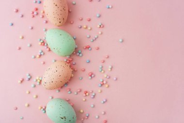 Pastel pembe ve yeşil renkte dört Paskalya yumurtası pembe bir yüzeye yerleştirilmiş, küçük, renkli şeker süslemeleriyle çevrili, neşeli bir atmosfer yaratıyor..