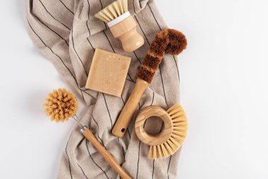 Çizgili bir kumaş üzerinde çeşitli doğal temizlik aletleri sergilenir. Düzenleme arasında fırçalar, sabun çubuğu ve çevre dostu tercihlerin altını çizen bir temizlik bezi yer alıyor..