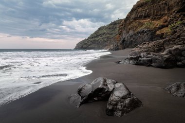 Önünde siyah bir kum ve büyük bir kaya olan kayalık bir sahil. Okyanus kıyısı, Tenerife, Kanarya Adaları ile seyahat ve turizm kavramı. 