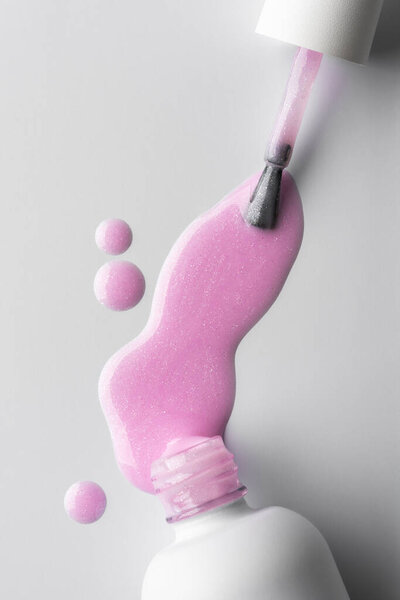 Close-up of pink shimmer nail polish spilling with glossy drops on a clean white surface. 