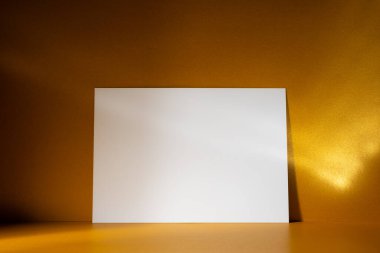 Işıldayan altın bir zemin üzerinde duran beyaz bir kağıt yaprağı ve güneş ışığı yansımaları. Minimalist ve zarif kurgu, modeller, davetiyeler veya lüks temalı tasarımlar için ideal.