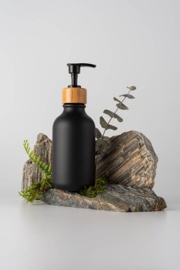 Taş podyumda yeşil yapraklı siyah şişe maketi. Doğal cilt bakımı ürünü markalaşma şablonu. Organik ve çevre dostu güzellik temaları için minimalist kompozisyon.