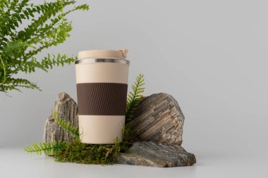 Kahverengi kabzalı ve kapaklı yeniden kullanılabilir kahve fincanı, yosun ve eğreltiotu ile kayaların üzerine yerleştirilmiş. Çevre dostu, sürdürülebilirlik, doğaya duyarlı yaşam tarzı ve markalaşma modellemeleri için minimum tasarım konsepti.
