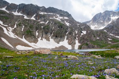 Dağ gölü ve kayalık dağlar. Kayaların ortasında berrak bir göl. Yeşil tarlalar, göl ve dağ sıraları karın altında.