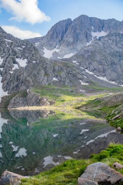 Dağ gölü ve kayalık dağlar. Kayaların ortasında berrak bir göl. Yeşil tarlalar, göl ve dağ sıraları karın altında.