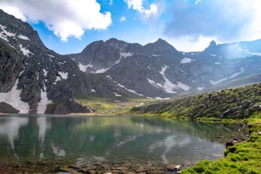 Dağ gölü ve kayalık dağlar. Kayaların ortasında berrak bir göl. Yeşil tarlalar, göl ve dağ sıraları karın altında.