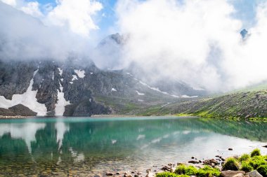 Dağ gölü ve kayalık dağlar. Kayaların ortasında berrak bir göl. Yeşil tarlalar, göl ve dağ sıraları karın altında.