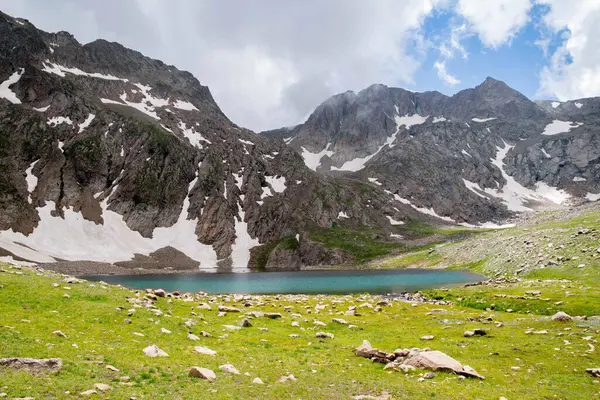 Dağ gölü ve kayalık dağlar. Kayaların ortasında berrak bir göl. Yeşil tarlalar, göl ve dağ sıraları karın altında.