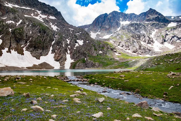 Dağ gölü ve kayalık dağlar. Kayaların ortasında berrak bir göl. Yeşil tarlalar, göl ve dağ sıraları karın altında.
