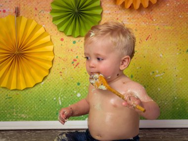 İlk doğum günü partisinde çekilen renkli görüntüler. Küçük sarışın bir çocuk doğum günü pastasını tahta kaşıklarla eziyor.