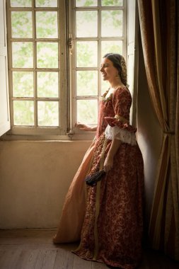 Otantik Rönesans kostümü ve başlık giymiş bir kadın ortaçağ şatosunun penceresinde dikiliyor.