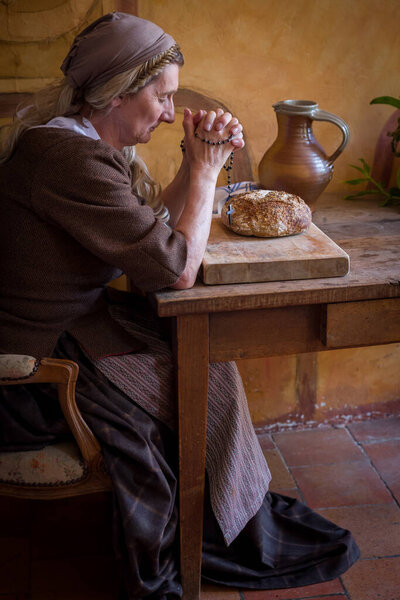 Woman in authentic peasant renaissance costume praying at a table. There is bread on the table.