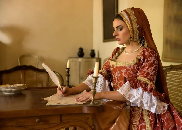 Woman wearing renaissance gown and French hood writing a letter at her table.