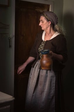 Ortaçağ kostümü giymiş bir kadın elinde bir sürahi taşla odaya giriyor.