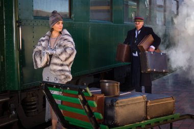 1927 model birinci sınıf bir tren vagonunun yanındaki platformda canlandırma sahnesinde 1920 'lerden kalma lüks bir bayan valizini bekliyor.
