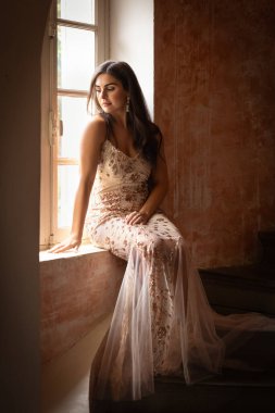 Altın pullu güzel bir esmerin pencere ışığında portresi. Antik bir sarmal merdivende elbise.