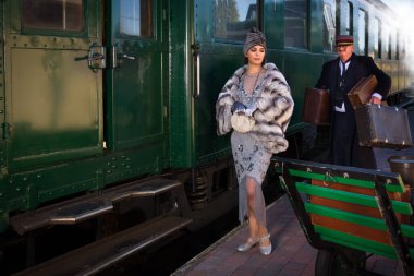 1927 model birinci sınıf bir tren vagonunun yanındaki platformda canlandırma sahnesinde 1920 'lerden kalma lüks bir bayan valizini bekliyor.