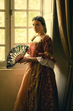 Otantik Rönesans kostümü ve başlık giymiş bir kadın ortaçağ şatosunun penceresinde dikiliyor.