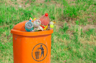 Turuncu çöp tenekesi çöp şişeleri ve kağıdıyla dolup taşarak yeşil çimlerin arka planında geri dönüşüm ve imha için gönderilmeyi bekliyor.
