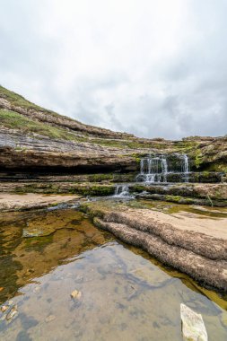 İspanya, Cantabria 'da yemyeşil yosunlarla kaplı katmanlı kaya oluşumlarının üzerinden akan resim gibi bir şelale. Doğa, seyahat ve manzara temaları için ideal.