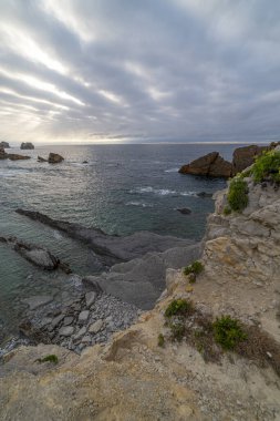 İspanya, Cantabria 'da dramatik gökyüzü, kayalık kıyı şeridi ve kıyı evleri içeren Playa de la Arna üzerinde güzel bir günbatımı. Deniz manzarası, seyahat ve doğa temaları için mükemmel.. 