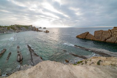 İspanya, Cantabria 'da dramatik gökyüzü, kayalık kıyı şeridi ve kıyı evleri içeren Playa de la Arna üzerinde güzel bir günbatımı. Deniz manzarası, seyahat ve doğa temaları için mükemmel.. 