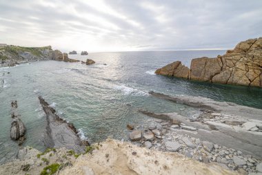 İspanya, Cantabria 'da dramatik gökyüzü, kayalık kıyı şeridi ve kıyı evleri içeren Playa de la Arna üzerinde güzel bir günbatımı. Deniz manzarası, seyahat ve doğa temaları için mükemmel.. 