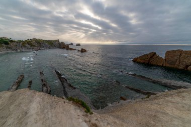 İspanya, Cantabria 'da dramatik gökyüzü, kayalık kıyı şeridi ve kıyı evleri içeren Playa de la Arna üzerinde güzel bir günbatımı. Deniz manzarası, seyahat ve doğa temaları için mükemmel.. 
