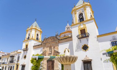 İspanya 'nın Ronda kentindeki Nuestra Senora de la Merced Kilisesi' nin göz kamaştırıcı cephesinde, parlak mavi gökyüzünün altında ikiz çan kuleleri ve merkezi bir çeşme bulunuyor. İspanyol mimarisi, dini simgeler ve kültür turizmi için mükemmel bir tema.