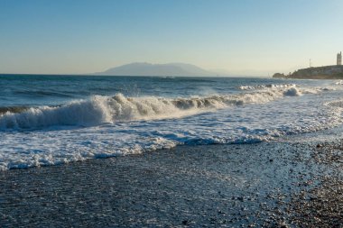 İspanya 'nın Malaga kıyıları boyunca güçlü dalgalar kopuyor ve ufukta uzak dağlar görünüyor. Kıyı enerjisi, doğa ve manzaralı deniz manzarası temaları için ideal