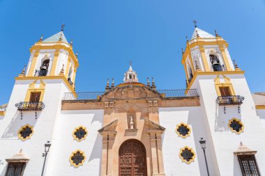 İspanya 'nın Ronda kentindeki Nuestra Senora de la Merced Kilisesi' nin göz kamaştırıcı cephesinde, parlak mavi gökyüzünün altında ikiz çan kuleleri ve merkezi bir çeşme bulunuyor. İspanyol mimarisi, dini simgeler ve kültür turizmi için mükemmel bir tema.